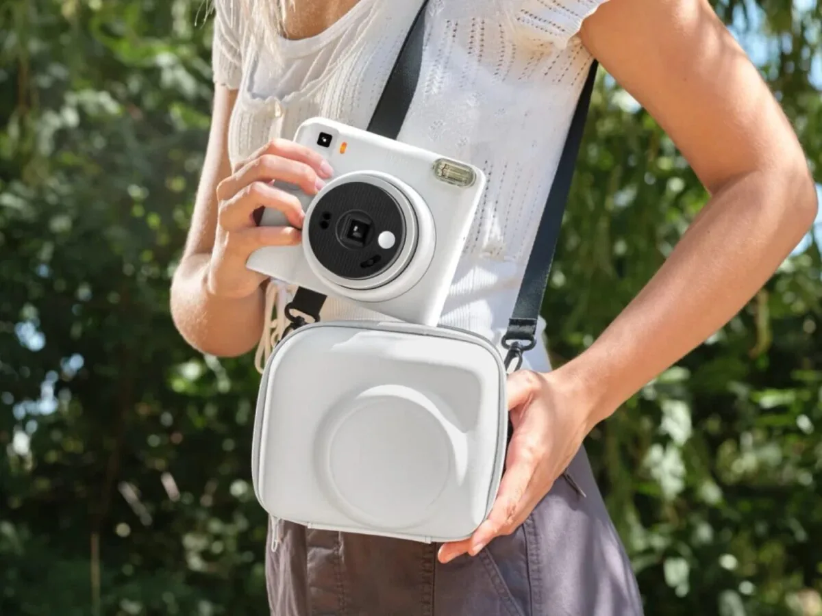 فردی با لباس تابستانی سفید و کیف دوشی سفید، دوربین چاپ سریع سفید Instax Square SQ2 را با بند مشکی روی گردن و دستان خود نگه داشته است.