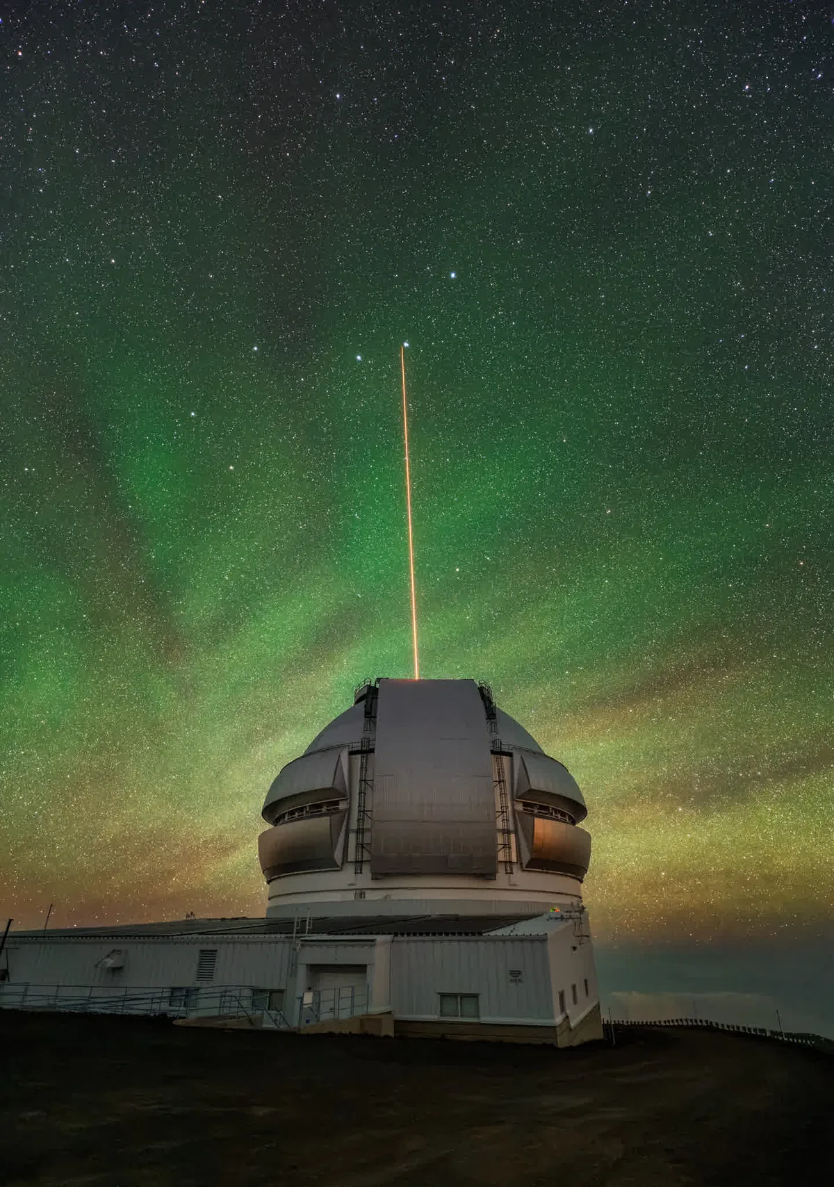 Night sky with stars and green aurora above a large observatory telescope; an orange beam rises from the dome into the sky.