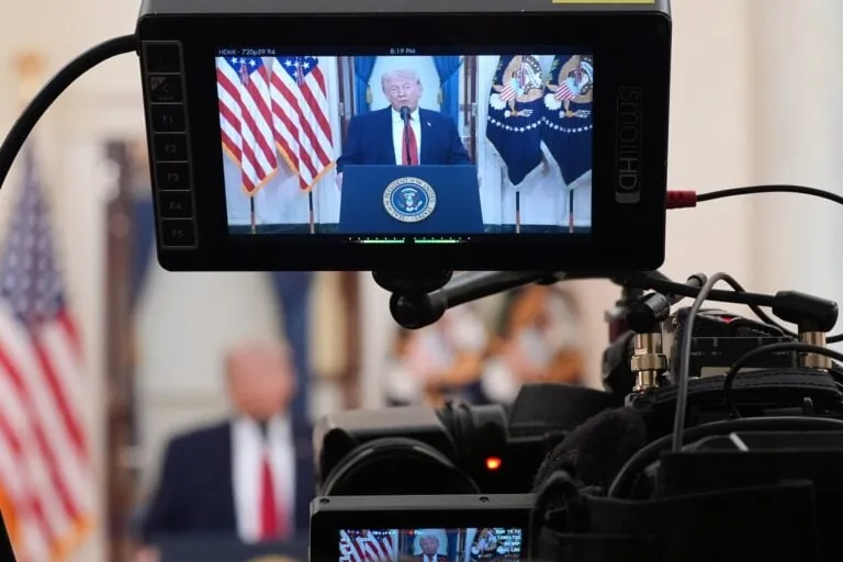 Monitor shows a man at a podium with the presidential seal, flanked by American flags, during a press address.