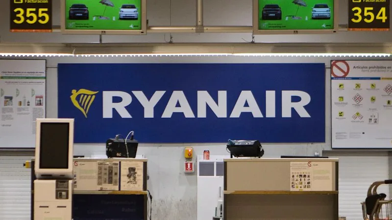 Airport check-in area with a large blue RYANAIR sign behind two service desks and overhead monitors showing flight numbers.