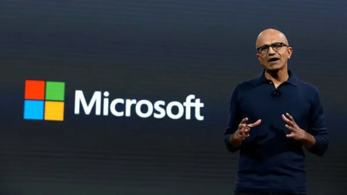 A bald man in a dark blue polo speaking on stage with a Microsoft logo behind him.