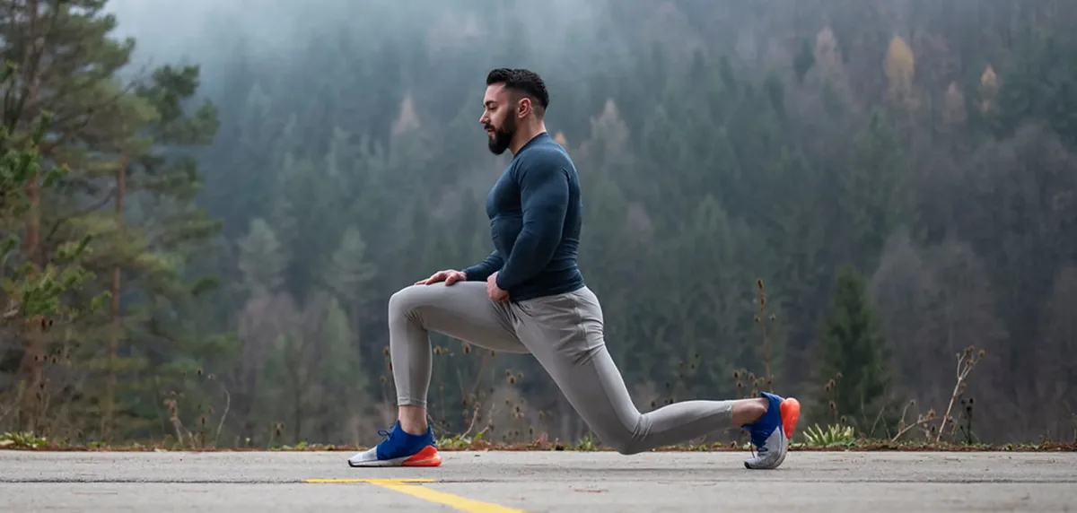 Man outdoors in a blue long-sleeve shirt and gray leggings performs a lunge on a paved path with a forest backdrop.