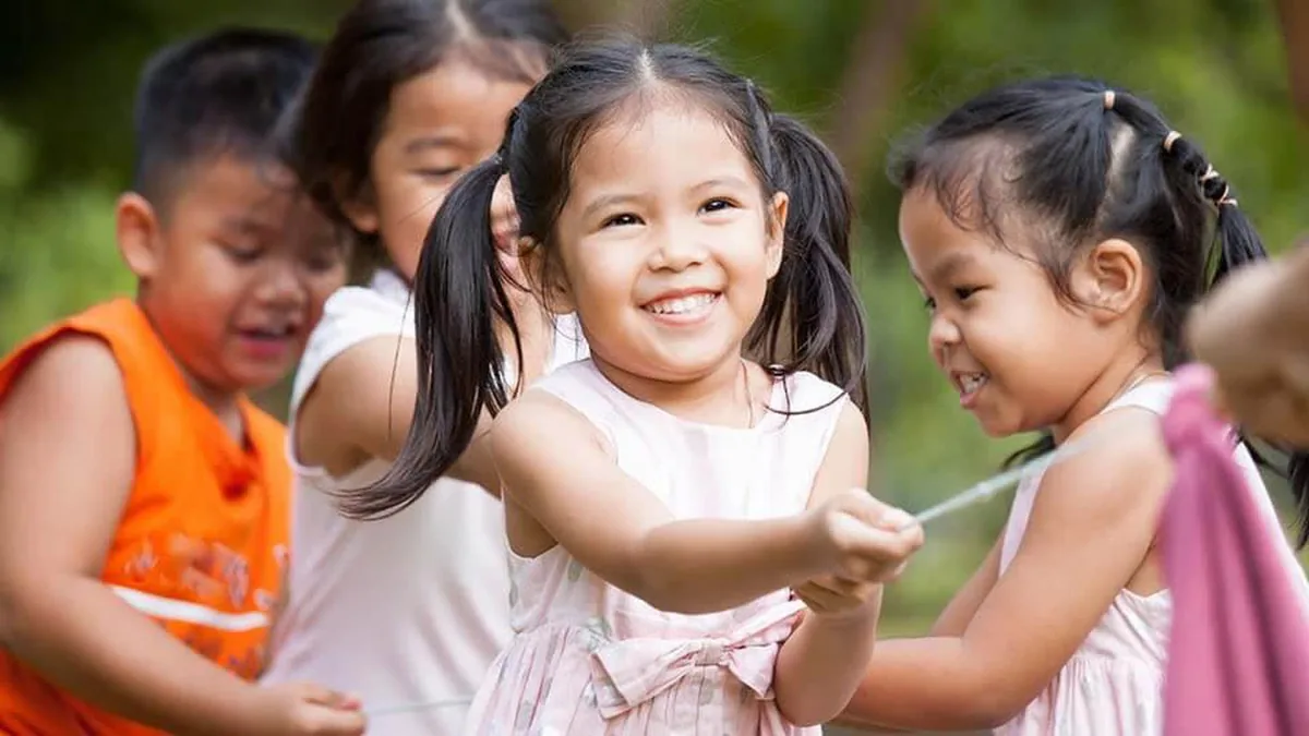 Smiling young girl with pigtails holding a jump rope while friends play outdoors in the background.