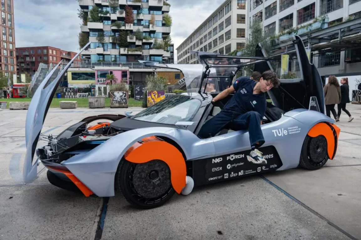 Futuristic silver and orange sports car with doors open, a person in navy overalls stepping into the cockpit in an urban plaza.