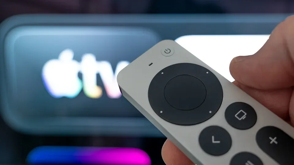 نمای نزدیک از دست فردی که ریموت کنترل نقره‌ای Apple TV را در دست گرفته و در پس‌زمینه محو، لوگوی درخشان Apple TV روی نمایشگر دیده می‌شود.